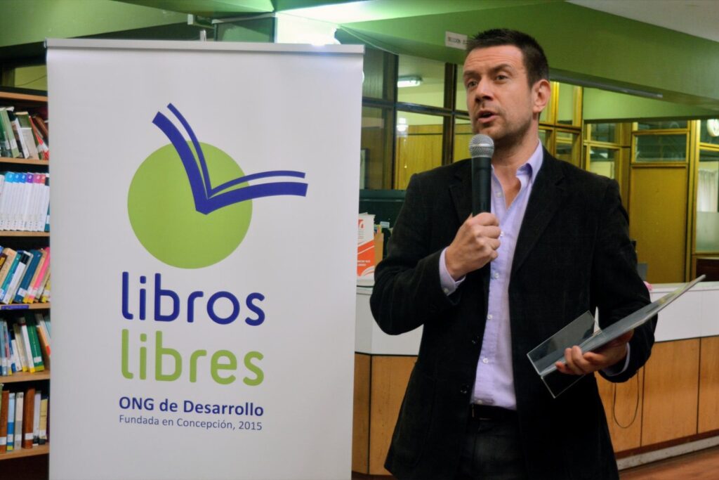 Fotografía de Mauricio Espina hablando al micrófono en una biblioteca, junto a un cartel de la ONG de Desarrollo “Libros Libres”, sosteniendo un documento en sus manos.