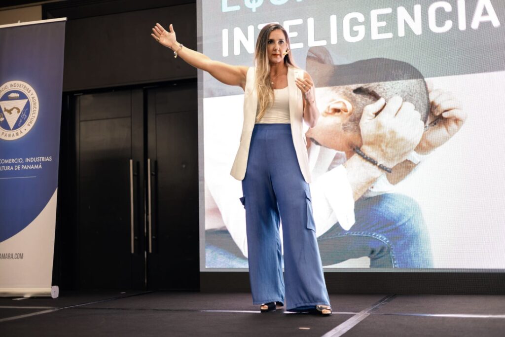 Fotografía de Meli Alemán durante una conferencia, captada mientras gesticula con el brazo extendido frente a una pantalla gigante que muestra la palabra "Inteligencia" y la imagen de un hombre con las manos en la cabeza. Escenario interior con iluminación profesional.