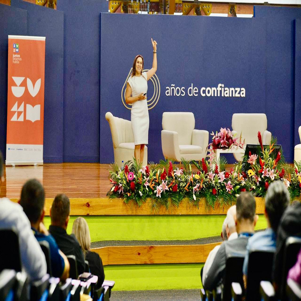 Fotografía de Nadia Vado en un auditorio, dando una conferencia desde el escenario decorado con flores y un fondo azul con el texto "años de confianza", vestida con atuendo formal blanco y dirigiéndose al público.