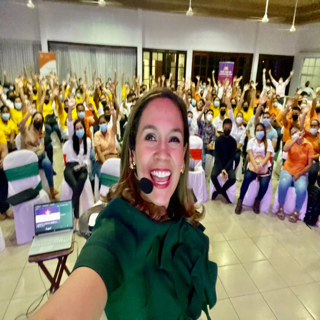 Fotografía de Nadia Vado en una conferencia, tomándose una selfie con micrófono de diadema mientras una audiencia entusiasta la acompaña al fondo levantando los brazos, en un salón cerrado con ambiente alegre.