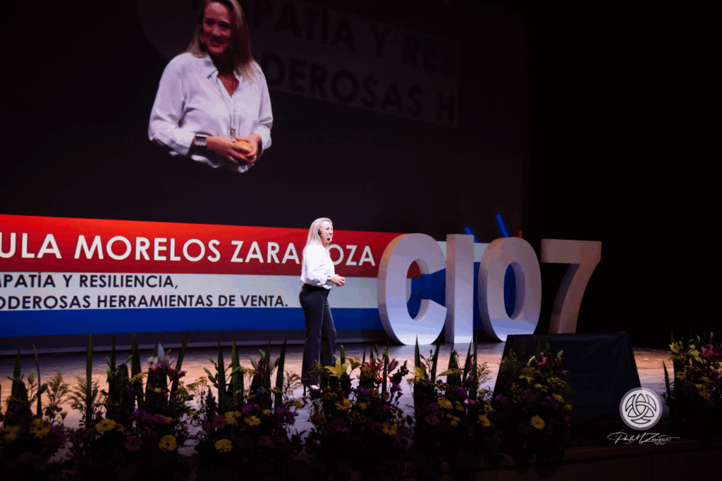 Fotografía de Paula Morelos Zaragoza impartiendo una conferencia en un auditorio, de pie sobre un escenario con flores al frente y una pantalla que muestra su nombre junto al tema “Empatía y resiliencia, poderosas herramientas de venta”.