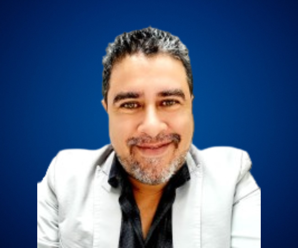 Fotografía de Rodrigo Quintanilla, con fondo azul sólido, luciendo saco claro y camisa oscura, sonriendo directamente a la cámara.