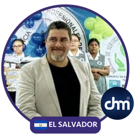 Fotografía de Rodrigo Quintanilla durante un evento en El Salvador, vistiendo saco claro y camiseta oscura, rodeado de personas con fondo decorado con globos y banners institucionales.