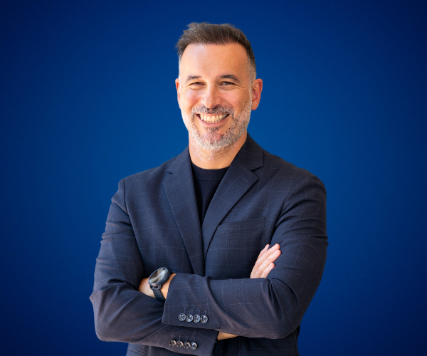 Fotografía de Rubén Montesinos, de pie frente a un fondo azul, sonriendo con los brazos cruzados y vistiendo un saco oscuro; imagen profesional con iluminación clara.