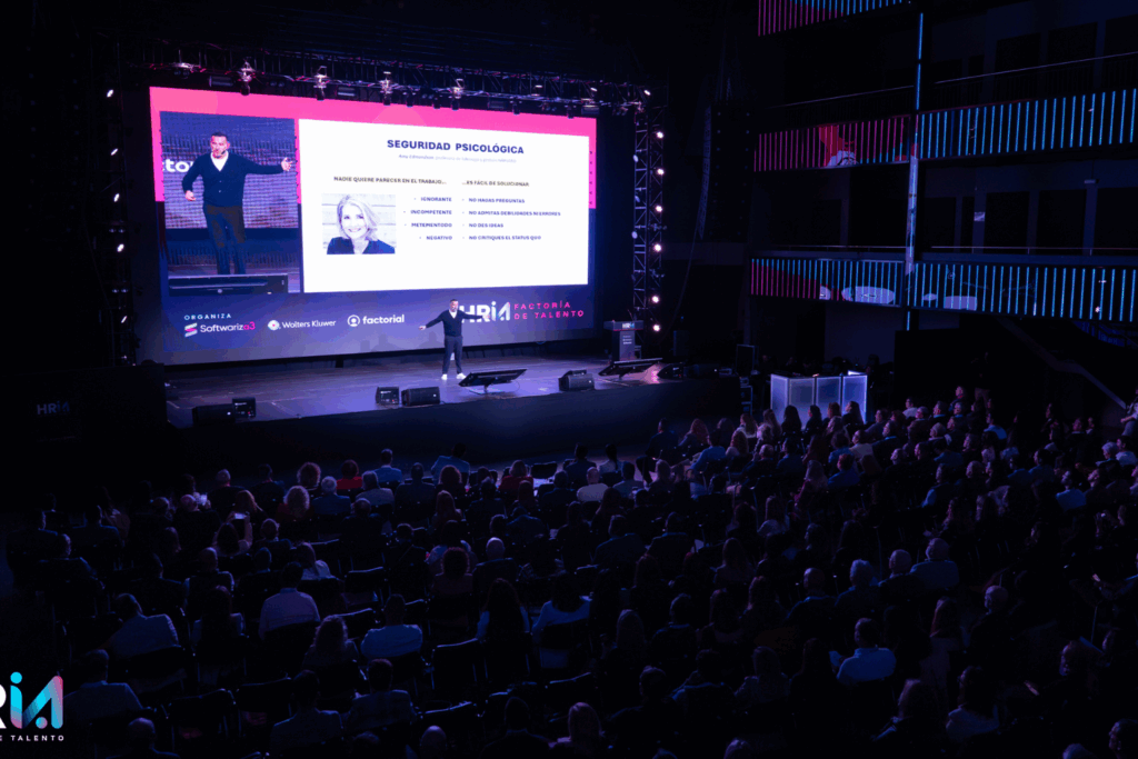 Fotografía de Rubén Montesinos dando una conferencia sobre seguridad psicológica ante un auditorio lleno, con gran pantalla proyectando contenido y luces escénicas.