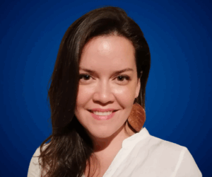 Fotografía de Simoné Oliva, conferencista, posando con una sonrisa cálida sobre fondo azul profesional, vestida con blusa blanca y aretes grandes color cobre.