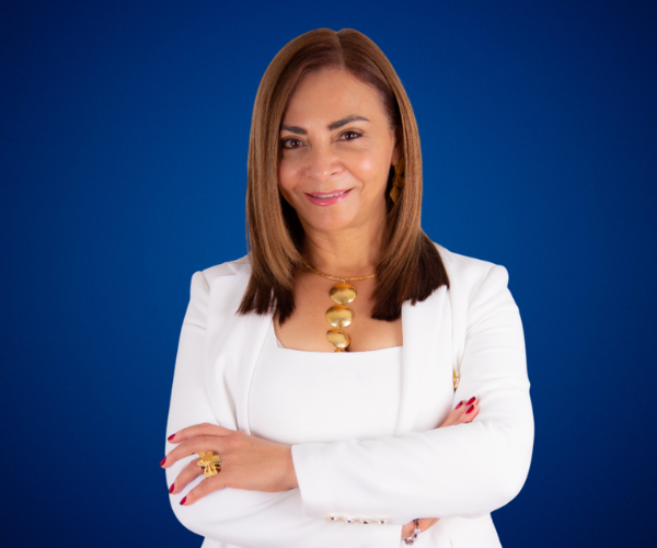 Fotografía de Sonia Rojas, posando con elegancia frente a un fondo azul, vestida con un traje blanco y accesorios dorados, transmitiendo confianza y profesionalismo.