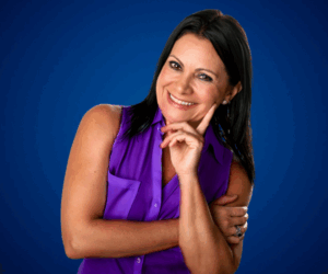 Fotografía de Yanira Puy, sonriente y con actitud cercana, vestida con blusa sin mangas color púrpura, sobre un fondo azul que resalta su presencia profesional.