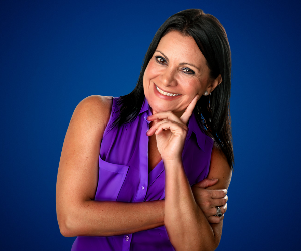 Fotografía de Yanira Puy, sonriente y con actitud cercana, vestida con blusa sin mangas color púrpura, sobre un fondo azul que resalta su presencia profesional.
