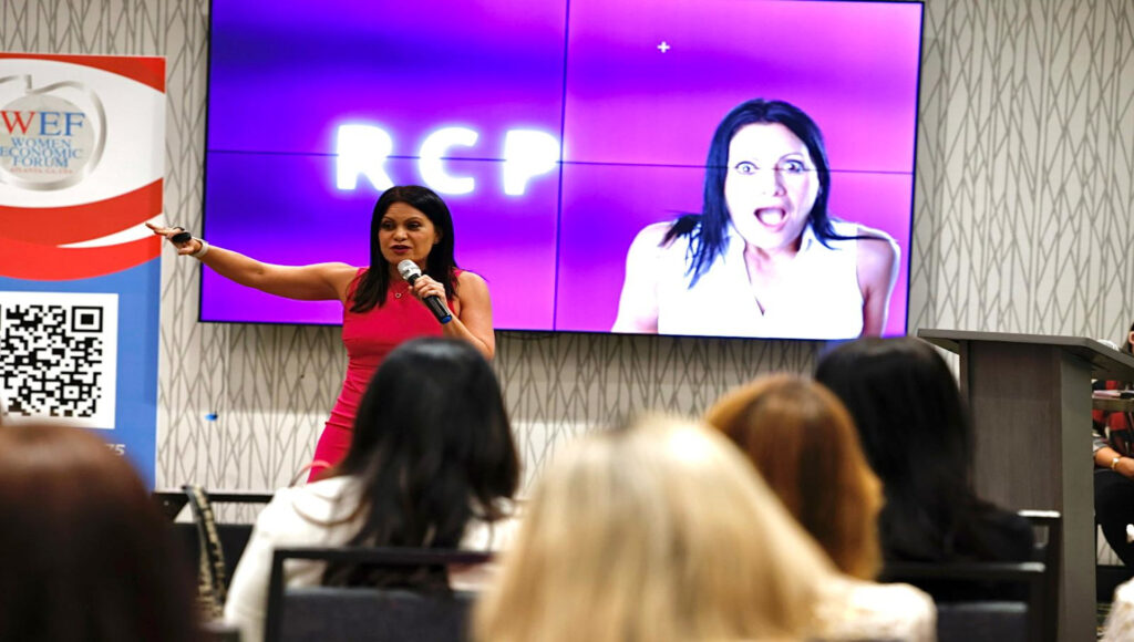 Fotografía de Yanira Puy impartiendo una conferencia en el Women Economic Forum, vestida con un elegante vestido fucsia, sosteniendo un micrófono frente a una audiencia atenta. En la pantalla aparece una imagen de ella con fondo púrpura y letras “RCP”.