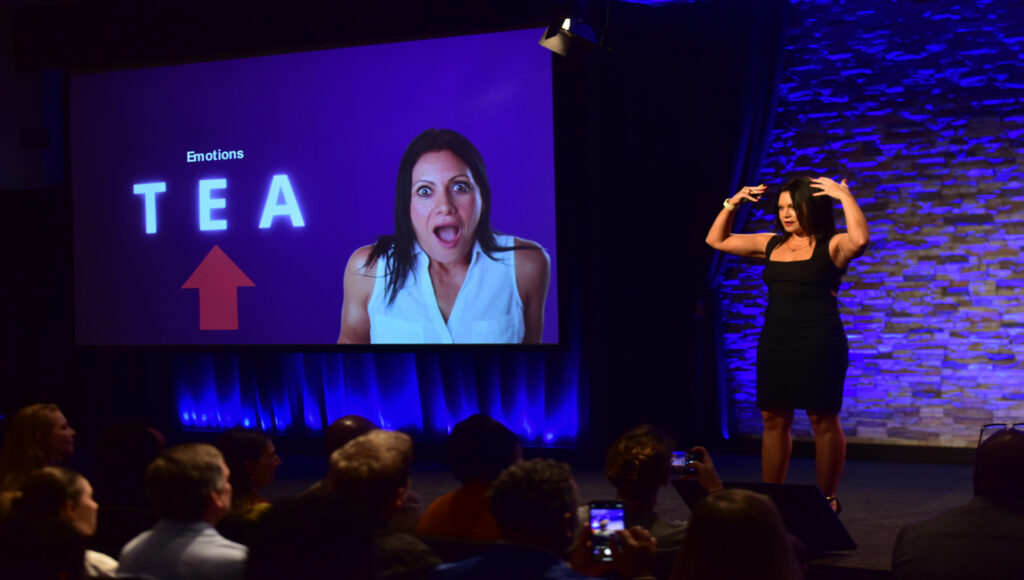 Fotografía de Yanira Puy en el escenario durante una conferencia, vestida con un elegante vestido negro, gesticulando con energía frente a una pantalla con la palabra “TEA” y la imagen de ella misma expresando emociones, ante un público atento.