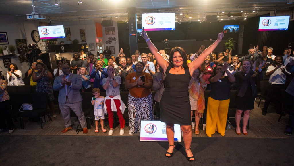 Fotografía de Yanira Puy en el escenario con los brazos en alto, recibiendo una ovación de pie de una audiencia entusiasta en un evento profesional.