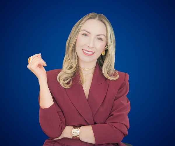 Fotografía de Yuly Giraldo, sonriente y elegante, vestida con un traje burdeos, sobre un fondo azul profesional. Su pose transmite confianza y calidez.