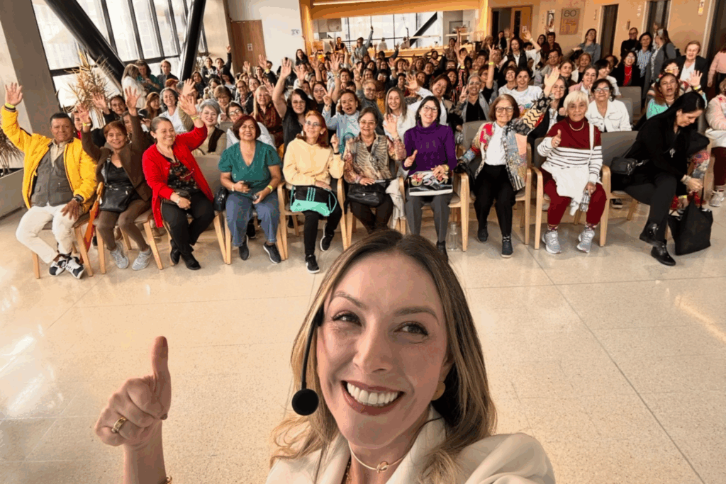 Fotografía de Yuly Giraldo en primer plano, sonriendo con micrófono de diadema, al frente de una audiencia entusiasta que levanta la mano en señal de participación dentro de un espacio iluminado y moderno.