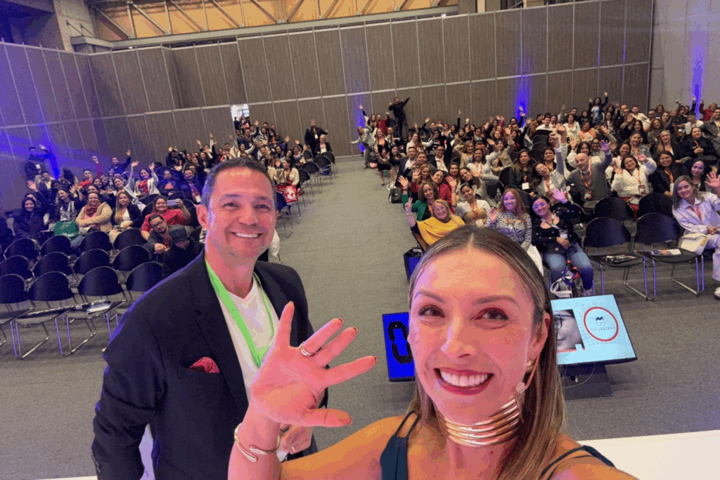 Fotografía de Yuly Giraldo junto a un conferencista invitado, ambos saludando sonrientes desde el escenario frente a una audiencia numerosa y entusiasta en un auditorio moderno con iluminación azul.