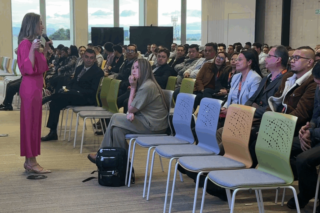 Fotografía de Yuly Giraldo de pie con micrófono en mano, vestida con traje fucsia, hablando frente a una audiencia empresarial atenta en un salón moderno con luz natural.