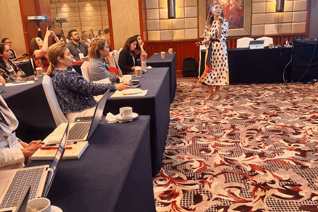 Fotografía de Yuly Giraldo dando una conferencia en salón de hotel, vestida con traje de puntos blanco con negro, frente a un grupo de profesionales atentos con laptops y cuadernos sobre mesas cubiertas con manteles oscuros.