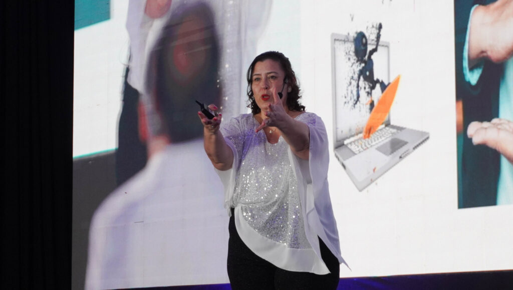 Fotografía de Emilse Silvestre en plena conferencia, gesticulando con energía frente a una pantalla gigante con imágenes digitales.