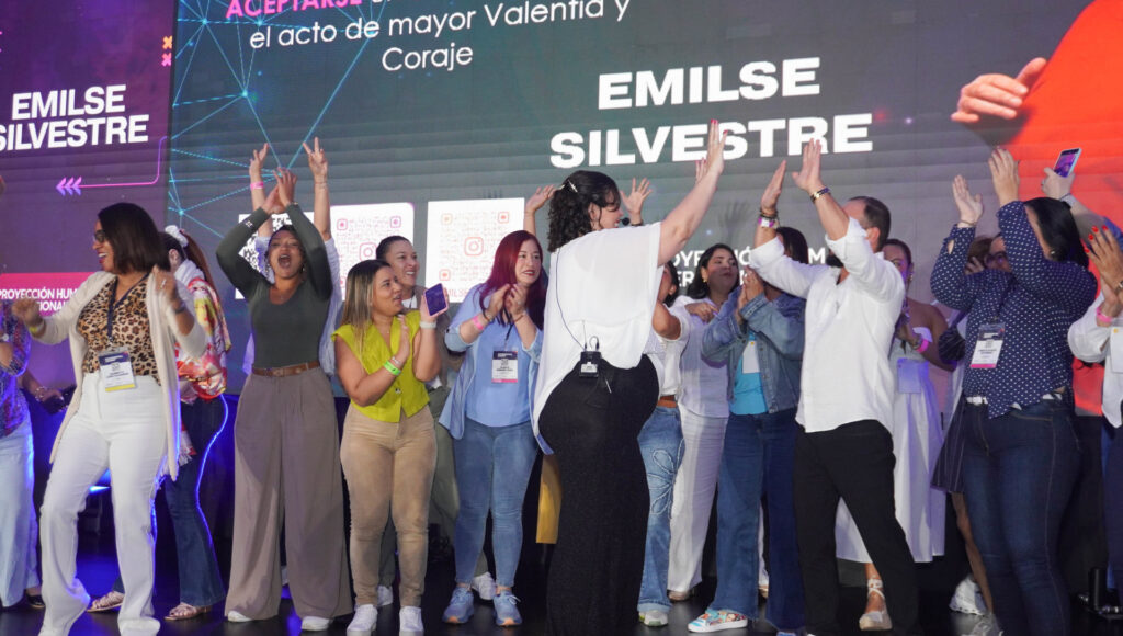 Fotografía de Emilse Silvestre en un escenario, interactuando con el público y celebrando junto a un grupo de personas entusiastas durante una conferencia.