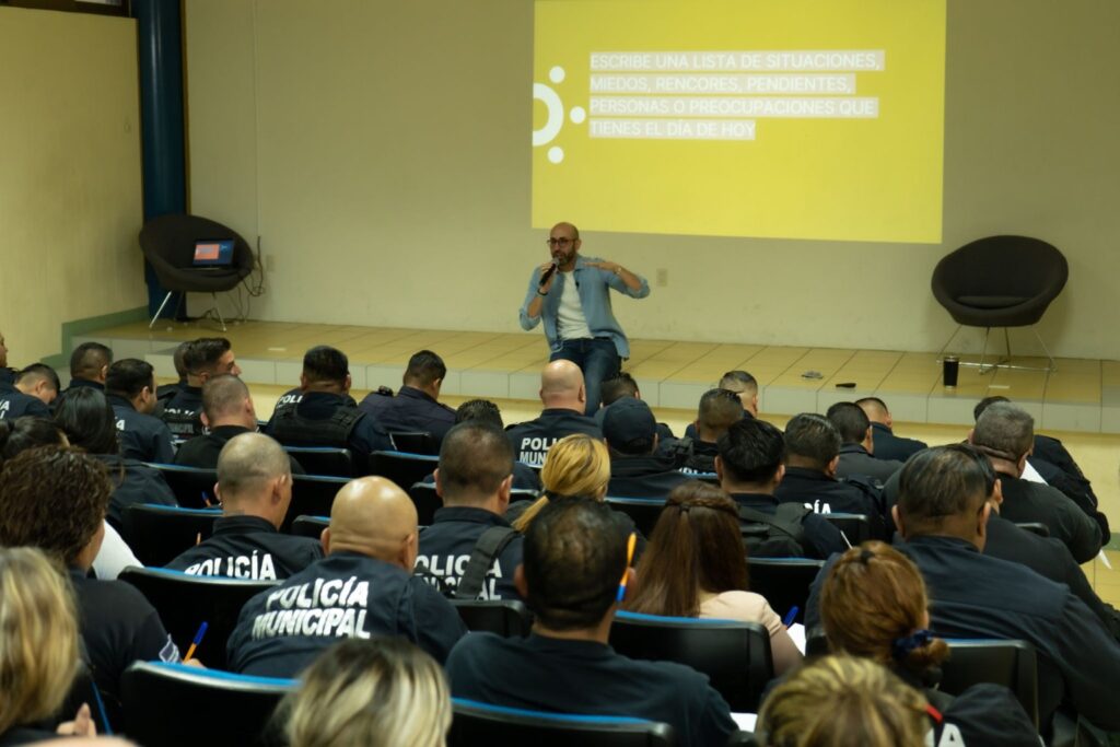 Fotografía de Anwar Buere impartiendo una conferencia a elementos de la Policía Municipal. Está sentado en el escenario con micrófono en mano, frente a un grupo atento de uniformados, con una diapositiva amarilla proyectada al fondo.