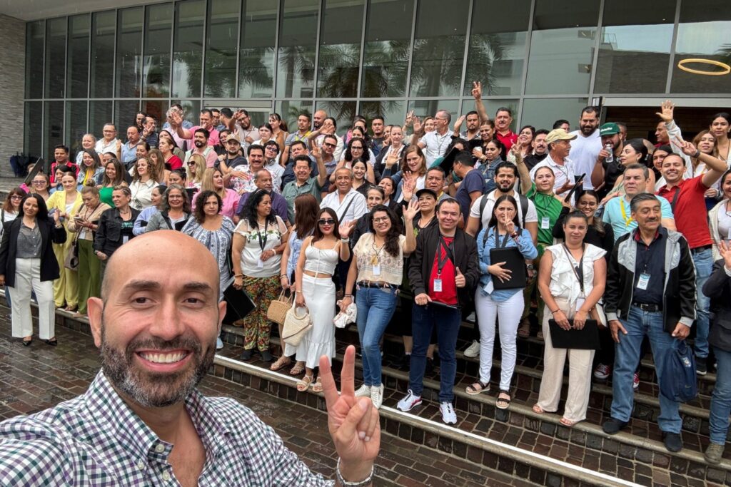 Fotografía de Anwar Buere tomando una selfie grupal con decenas de personas sonrientes sobre unas escaleras, todos en ambiente de camaradería frente a un edificio moderno de vidrio.