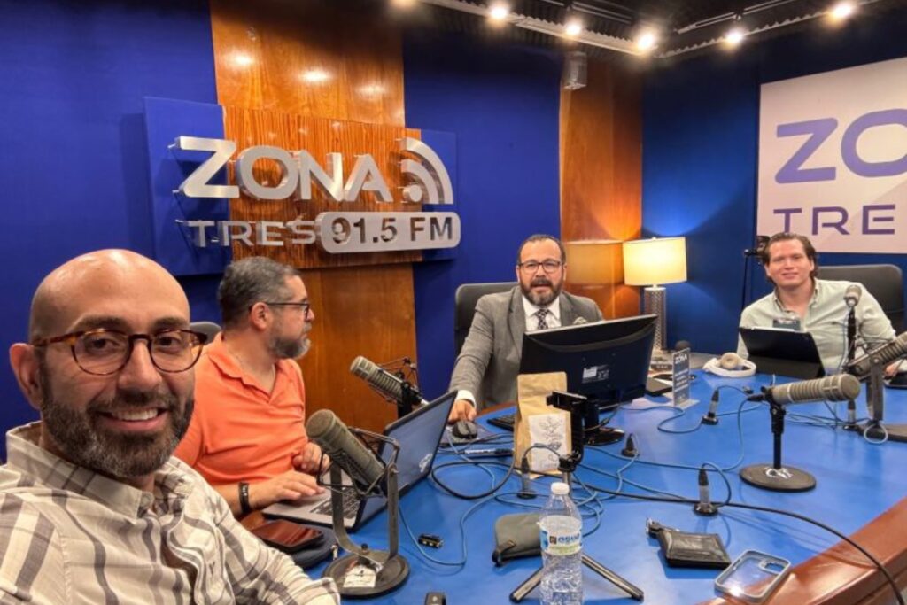 Fotografía de Anwar Buere en cabina de radio durante una transmisión en vivo en Zona Tres 91.5 FM, acompañado de tres conductores, rodeados de micrófonos, laptops y equipo de grabación profesional.