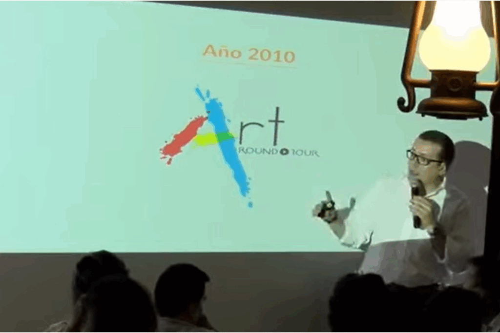 Fotografía de Gustavo De La Vega, conferencista, durante una presentación en sala interior, proyectando una diapositiva con el título "Año 2010" y el logo colorido de "Art Around Tour", mientras habla al público con micrófono en mano.
