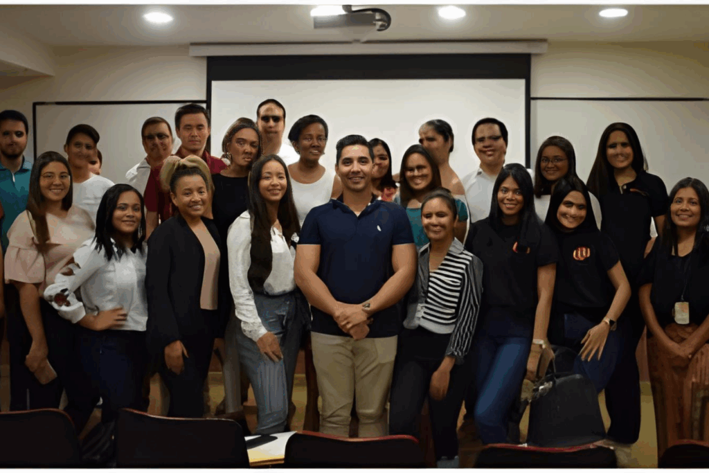 Fotografía de Gustavo De La Vega, conferencista, posando con un grupo de asistentes en un auditorio, después de una conferencia, frente a una pantalla de proyección.