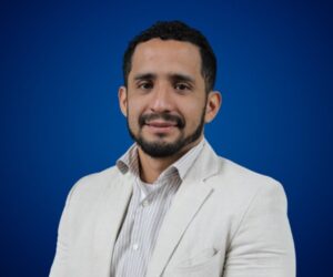 Fotografía de Jorge Moreno, conferencista internacional, posando con una leve sonrisa, luciendo saco beige claro y camisa de rayas, sobre un fondo azul profesional.