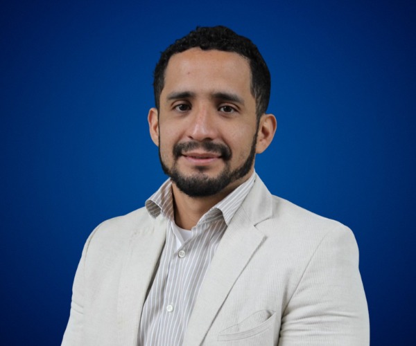 Fotografía de Jorge Moreno, conferencista internacional, posando con una leve sonrisa, luciendo saco beige claro y camisa de rayas, sobre un fondo azul profesional.