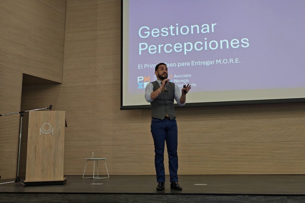 Fotografía de Jorge Moreno impartiendo una conferencia titulada “Gestionar Percepciones”, de pie en un escenario con micrófono en mano, vestido con chaleco gris y pantalón azul, frente a una pantalla de presentación.
