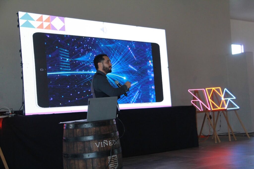 Fotografía de Jorge Moreno impartiendo una conferencia sobre transformación digital, de pie frente a una pantalla con gráficos tecnológicos en tonos azules, sosteniendo un micrófono y señalando la proyección.
