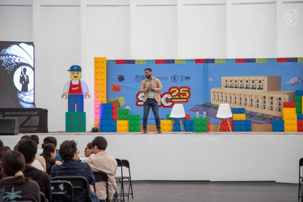 Fotografía de Jorge Moreno ofreciendo una conferencia frente a una audiencia, en un escenario decorado con piezas gigantes de Lego y un fondo colorido con temática universitaria.