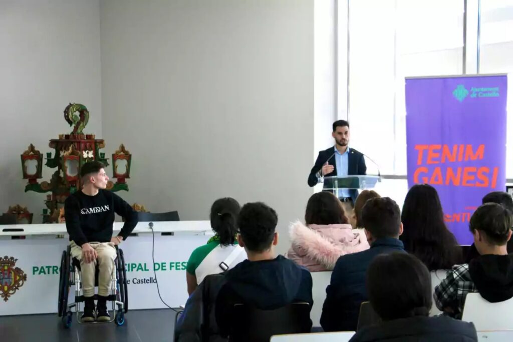 Fotografía de Carlos Tatay durante una charla institucional, sentado en su silla de ruedas frente a un grupo de jóvenes, mientras otro conferencista habla desde un atril junto a un cartel del Ayuntamiento de Castelló.