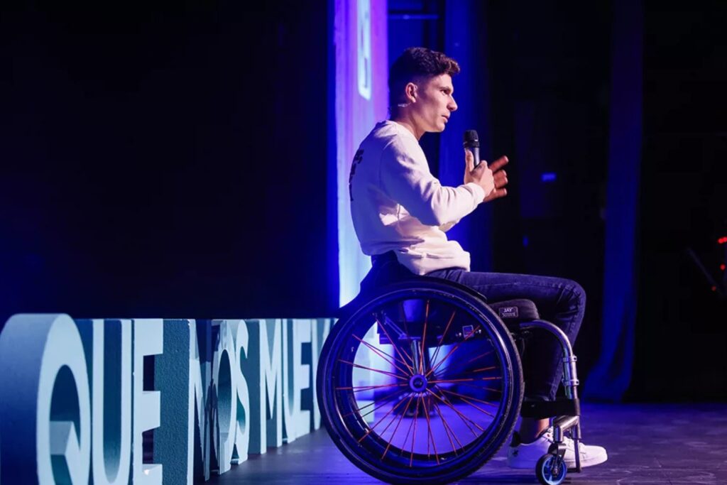 Fotografía de Carlos Tatay en plena conferencia, sentado en su silla de ruedas sobre un escenario iluminado con luces azules, mientras sostiene un micrófono y gesticula con energía.