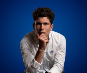Fotografía de Carlos Tatay, conferencista español, posando en actitud reflexiva con la mano en el mentón. Viste camisa blanca y se encuentra sobre un fondo azul sólido que resalta su expresión concentrada.