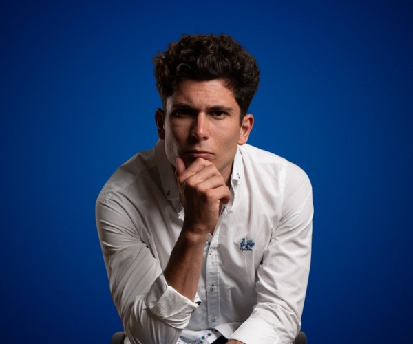 Fotografía de Carlos Tatay, conferencista español, posando en actitud reflexiva con la mano en el mentón. Viste camisa blanca y se encuentra sobre un fondo azul sólido que resalta su expresión concentrada.