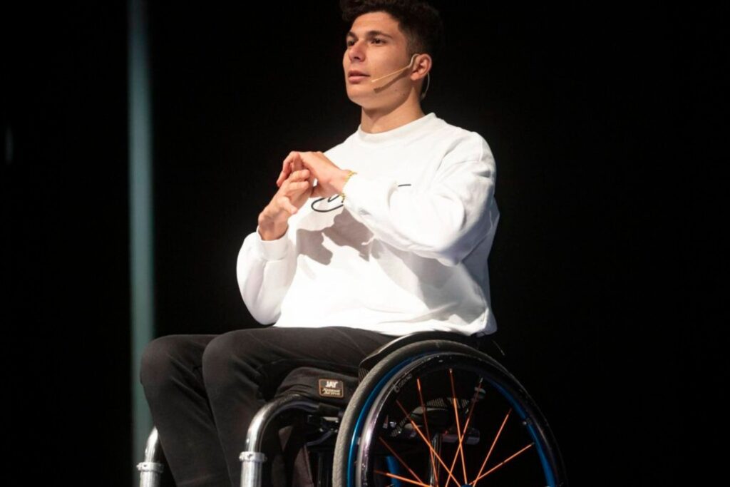 Fotografía de Carlos Tatay, conferencista de resiliencia, en el escenario con micrófono de diadema, sentado en su silla de ruedas deportiva, vestido con sudadera blanca, bajo una iluminación focal que destaca su expresión decidida.