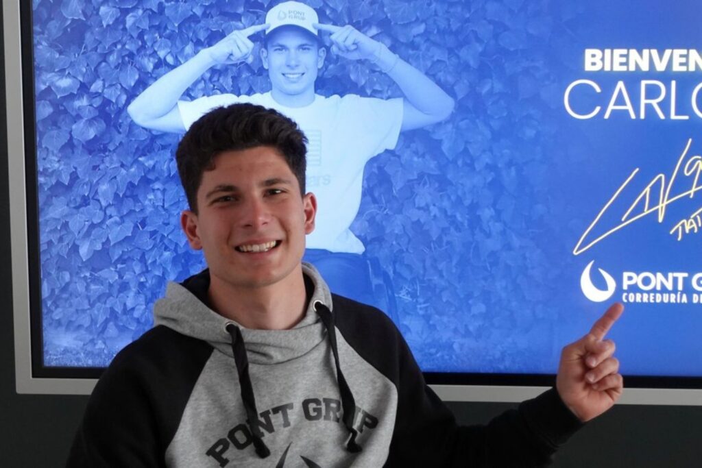 Fotografía de Carlos Tatay sonriendo y señalando una pantalla con su nombre y una imagen promocional de fondo, en un ambiente corporativo y juvenil.