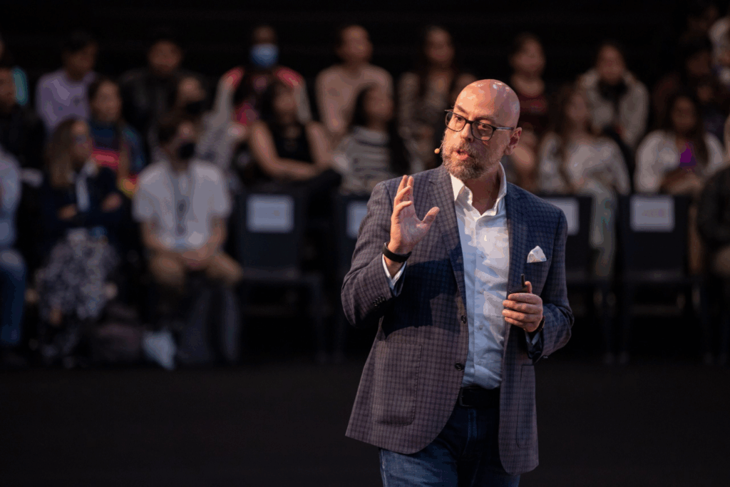 Fotografía de Sal Barrena en plena conferencia, vestido con saco a cuadros y camisa blanca, hablando con expresividad ante una audiencia atenta en un auditorio.