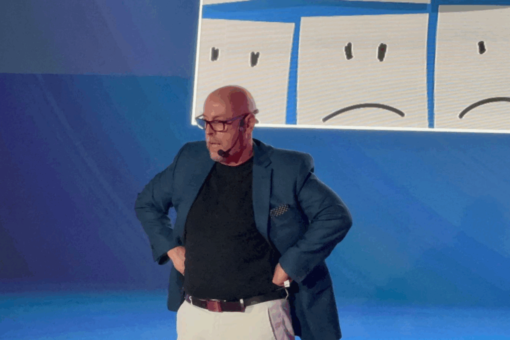 Fotografía de Sal Barrena durante una conferencia, con chaqueta azul, camiseta negra y pantalón claro, usando micrófono de diadema frente a una pantalla con ilustraciones de rostros tristes sobre un fondo azul.