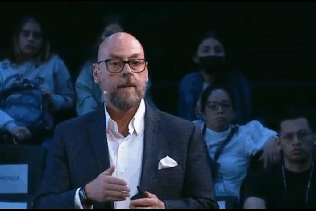 Fotografía de Sal Barrena durante una conferencia, vestido con saco oscuro y camisa blanca, usando micrófono de diadema mientras se dirige al público con gesto enfático y fondo de asistentes atentos.