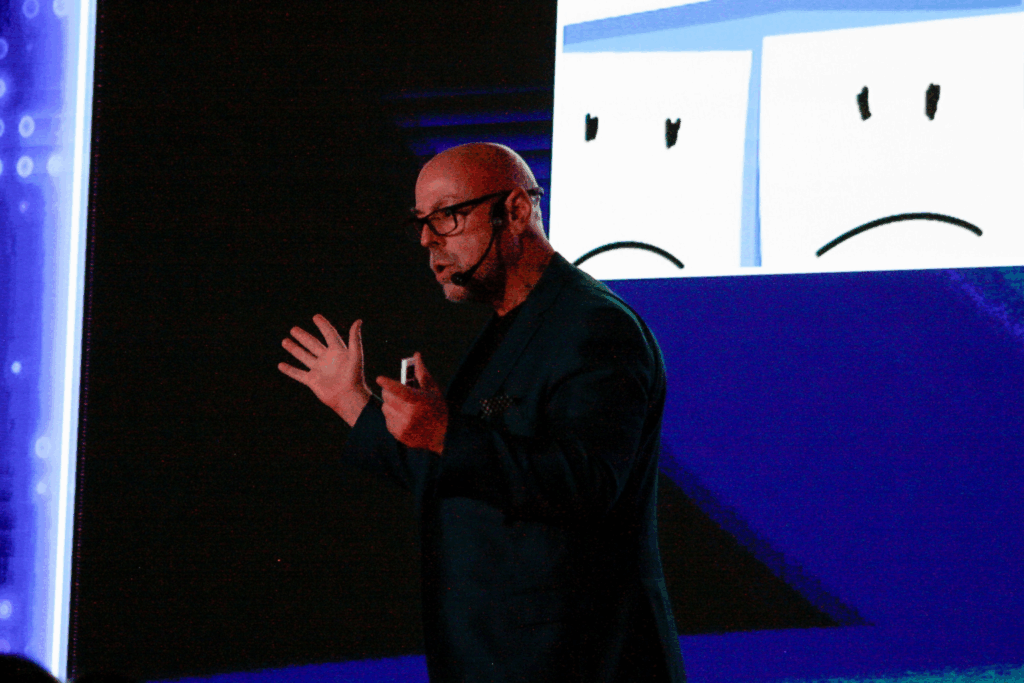Fotografía de Sal Barrena durante una conferencia, con micrófono de diadema, chaqueta oscura y fondo azul con ilustraciones de rostros tristes, gesticulando con energía mientras explica un concepto.