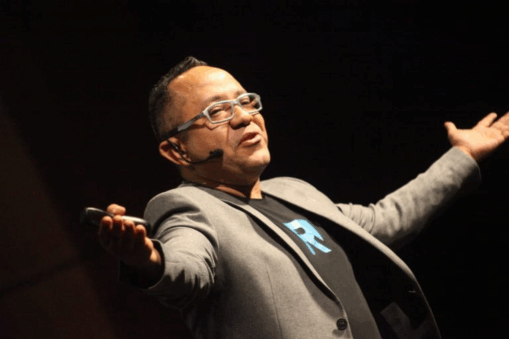 Fotografía de Raúl Camacho Rodríguez en plena conferencia, con micrófono de diadema, brazos extendidos y expresión entusiasta, sobre fondo oscuro.