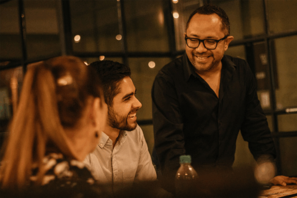 Fotografía de Raúl Camacho Rodríguez sonriendo mientras conversa con dos personas en un ambiente cálido e informal, con iluminación tenue.
