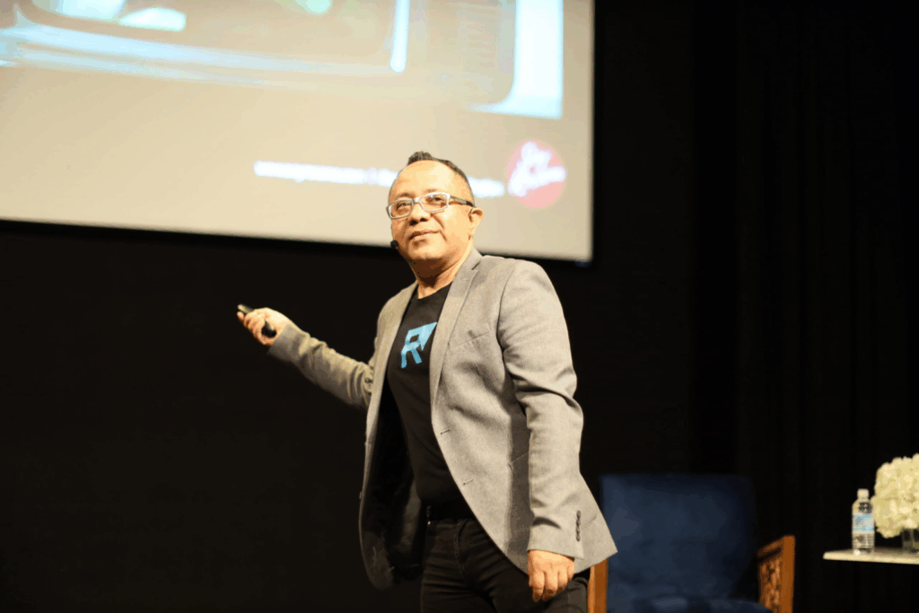 Fotografía de Raúl Camacho Rodríguez señalando una pantalla durante su conferencia, vestido con chaqueta gris y camiseta negra, en un escenario profesional.