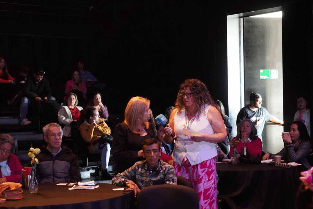 Fotografía de Geraldine Manríquez en una sala oscura de evento, entrevistando a una mujer con micrófono en mano y tarjetas; público atento sentado alrededor en mesas redondas.