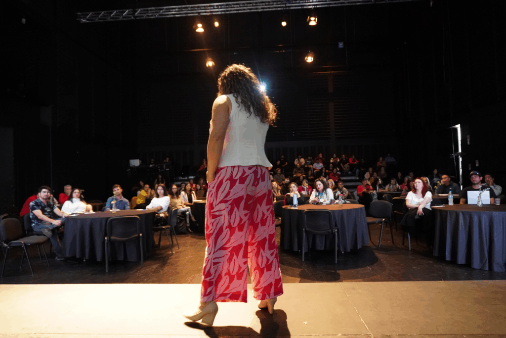 Fotografía de Geraldine Manríquez vista desde atrás sobre un escenario, vestida con pantalón fucsia estampado y blusa blanca, frente a una audiencia atenta en un salón oscuro con mesas redondas.