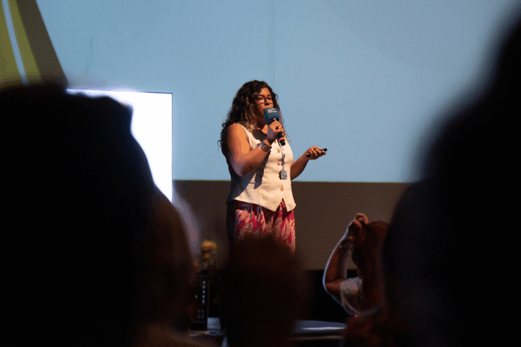 Fotografía de Geraldine Manríquez sobre un escenario, hablando al micrófono frente a una audiencia; viste chaleco blanco y pantalón fucsia estampado, con fondo de proyección iluminado.