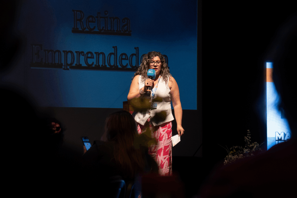 Fotografía de Geraldine Manríquez sobre un escenario oscuro, hablando al micrófono frente a una pantalla que proyecta el texto "Retina Emprendedora"; viste chaleco blanco y pantalón fucsia con estampado.